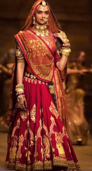 A woman dressed as an Indian bride stands gracefully in a dimly lit, royal-style setting, wearing a deep red lehenga with intricate gold embroidery, a matching dupatta draped over her head, and heavy traditional gold jewelry including a maang tikka, nose ring, necklaces, bangles, and earrings.